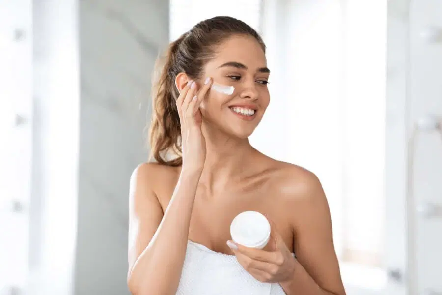 Smiling woman applying moisturizer to her cheek - What Are The 5 Basic Of Skin Care? 2026 - Zicail Smiling woman applying moisturizer to her cheek - What Are The 5 Basic Of Skin Care? 2026 - Zicail