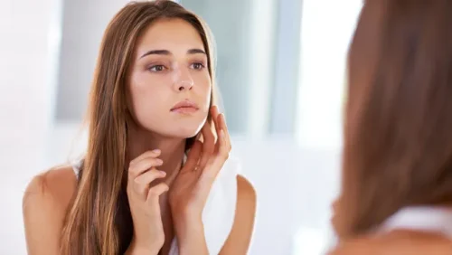 若い女性が鏡で自分の顔のシミをチェックしている - 透明感のある肌を手に入れる方法：健康的で輝く肌のためのヒント 2026 - Zicail